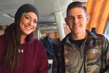 woman in maroon hoodie and beanie smiles next to man in camo jacket in the hangar