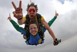 instructor in red sunglasses and yellow shirt shows a peace sign while jumping with young woman in blue jumpsuit