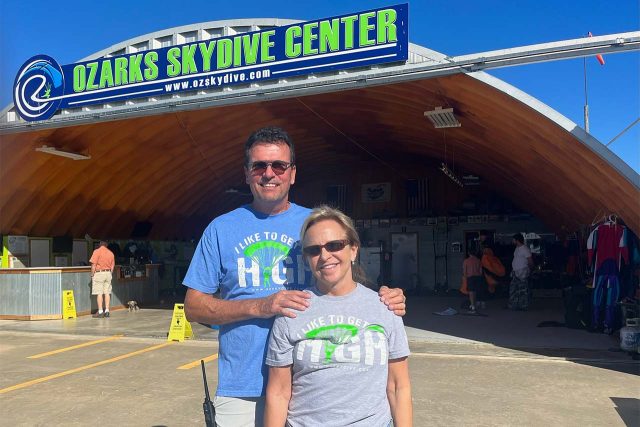 Bryan and Lyn Wolford stand in front of hangar and OZARKS Skydive Center sign