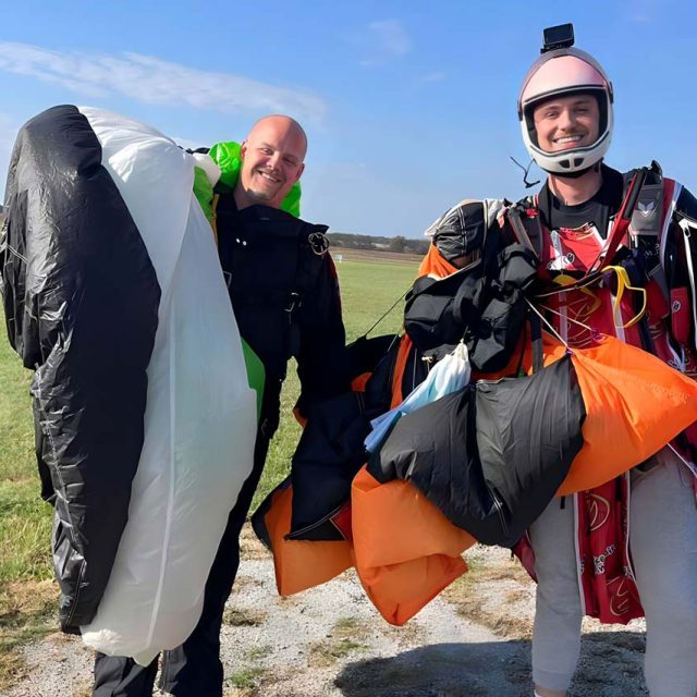 two instructors smile in the landing area with parachutes hanging over their shoulders