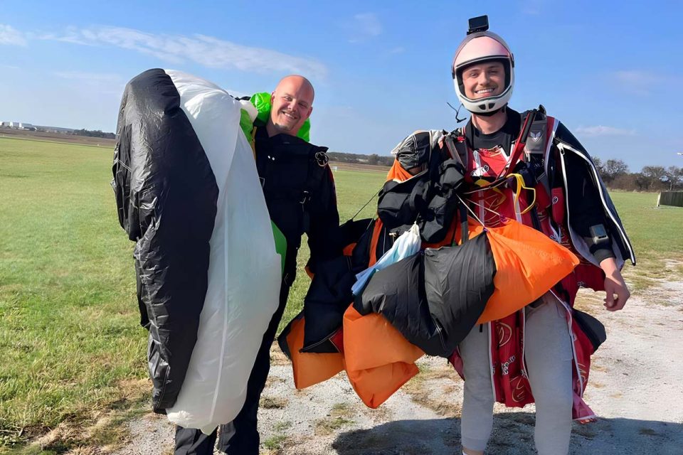two instructors smile in the landing area with parachutes hanging over their shoulders