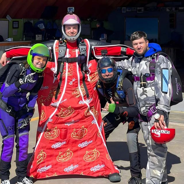 man wearing red wingsuit puts his arm around 3 other experienced jumpers