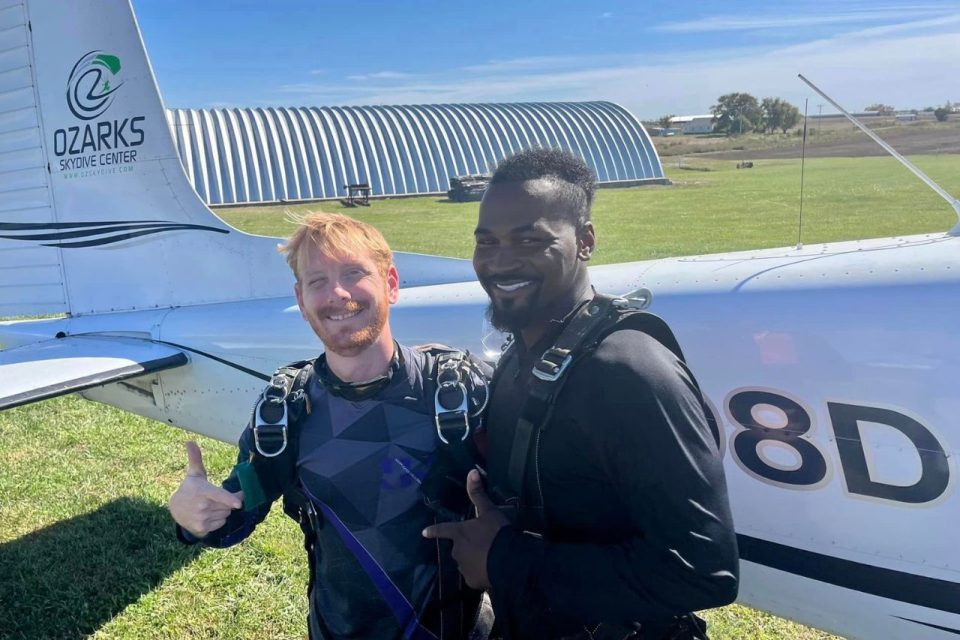 red-haired instructor and african-american man pose in front of aircraft before skydiving