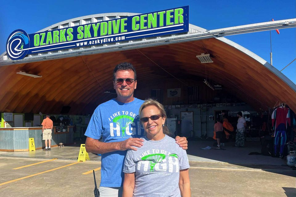 Bryan and Lyn Wolford stand in front of hangar and OZARKS Skydive Center sign