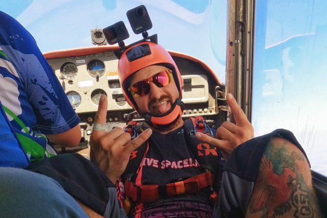 videographer in red helmet poses for a picture with tongue out while the plane climbs to altitude