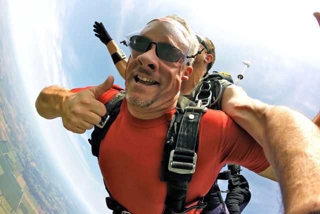 Man wearing red shirt and sunglasses gives a thumbs up in skydiving freefall