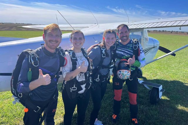two young women in harnesses give a thumbs up before getting into jump plane