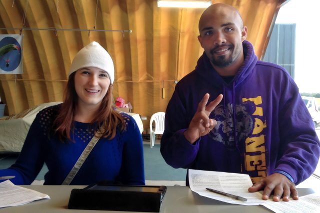 young woman in white beanie stands beside man in purple hoodie while checking in at manifest for their skydives